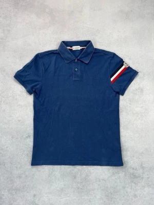 Blå pikétröja från Moncler - Snygg blå pikétröja från Moncler med klassisk krage och korta ärmar. På ena ärmen finns en röd, vit och svart randig detalj samt Moncler-logga. Perfekt för en stilren och sportig look.