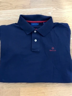 pikétröja från GANT - Klassisk mörkblå pikétröja från GANT med korta ärmar och krage. Tröjan har två knappar framtill och röd broderad logga på bröstet. Bra skick och passar till allt. Storlek: S