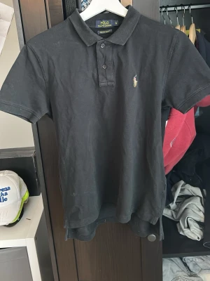Svart pikétröja från Ralph Lauren - Klassisk svart pikétröja från Ralph Lauren med gul broderad logga på bröstet. Modellen är Custom Slim Fit och har krage, korta ärmar och knäppning med tre knappar. Perfekt för en stilren och avslappnad look.