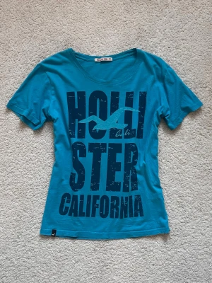 Hollister t-shirt - Blå 2000’s coded t-shirt från Hollister med stort tryck 'Hollister California'. T-shirten har rund halsringning och korta ärmar. Har använt få gånger och aldrig märkt det lilla hålet, syns inte på starka färgen.