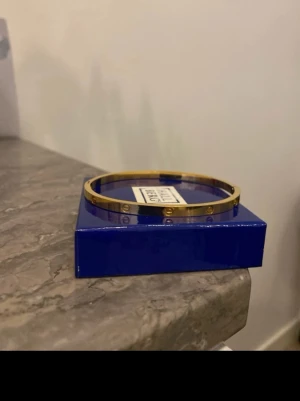 Guldfärgat armband  - Snyggt och stilrent armband i guldfärgad metall . Armbandet har en tunn, rund form och är dekorerat med de ikoniska skruvdetaljerna runt om. Perfekt accessoar för dig som gillar klassisk och lyxig design.