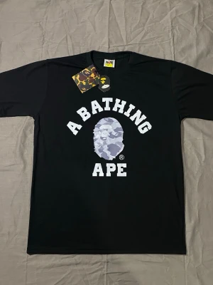 BAPE TSHIRT - Svart t-shirt från A Bathing Ape med stort vitt tryck och ikoniskt camo-mönstrat apansikte på bröstet. Klassisk passform och rund hals. Perfekt för dig som gillar streetwear och vill sticka ut med en ikonisk design.
