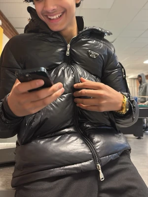 Svart pufferjacka från Prada - Snygg svart pufferjacka från Prada med glansig finish och klassisk logga på bröstet. Jackan har dragkedja framtill, hög krage och två sidofickor med dragkedja. Perfekt för kalla vinterdagar och ger en modern streetwear-look.FREE BISHAAR HORGAHBA SÅLD