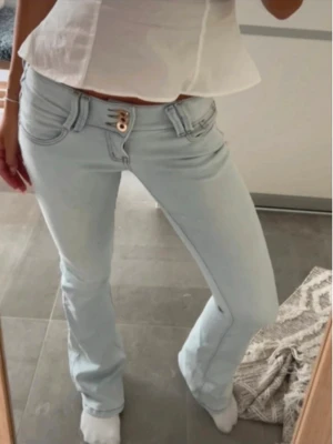 Ljusblå bootcut jeans lowwaist - Säljer ett par ljusblå bootcut jeans med dubbla guldiga knappar i midjan och klassiska fickor fram och bak. Jeansen har en snygg, figurnära passform upptill och lätt utsvängda ben. Perfekta för dig som gillar en trendig och avslappnad stil.