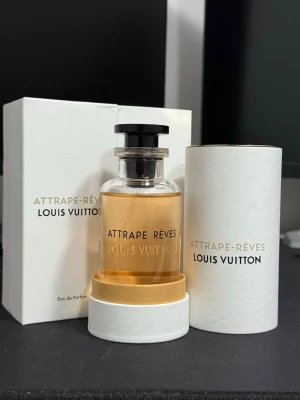 Louis Vuitton Attrape-Rêves parfym 100ml - Lyxig parfym från Louis Vuitton, Attrape-Rêves, i elegant glasflaska med svart lock och guldfärgad detalj. Doften kommer i en stilren vit förpackning med guld- och svart text. Flaskan rymmer 100 ml Eau de Parfum och utstrålar exklusivitet och modern design.
