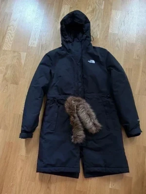 Svart parkas från The North Face - Svart parkas från The North Face med huva och avtagbar fuskpälsdetalj. Jackan är lång, har dragsko i midjan och logga på bröstet. Perfekt för kalla dagar med sin rejäla och stilrena design.