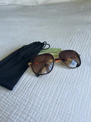 Guldiga solglasögon från Lacoste - Snygga solglasögon från Lacoste med fyrkantiga bruna glas och guldfärgade metallbågar. Klassisk design med diskret Lacoste-logga på ena glaset. Kommer med svart fodral och putsduk. Perfekta för soliga dagar och en stilren look.