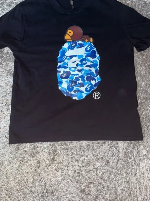 Svart BAPE t-shirt med blått camo - Svart t-shirt från BAPE med ikoniskt blått camo-tryck och apansikte på bröstet. Klassisk rund hals och korta ärmar. Tillverkad i bomull och har märkets signaturtaggar kvar. Perfekt för dig som gillar streetwear och unika prints.