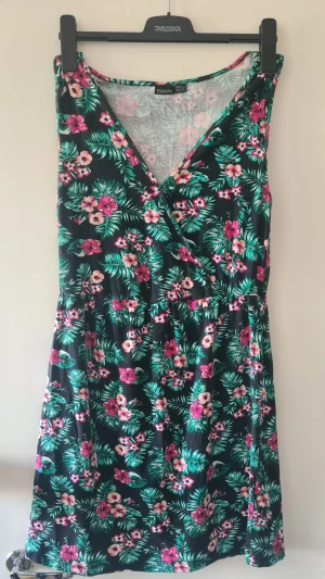 Blommig omlottklänning från Esmara - Snygg kort blommig klänning från Esmara med omlott och v-ringning. Klänningen har ett tropiskt mönster i grönt, rosa och vitt på svart botten. Ärmlös modell som går över knäet.