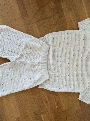 Vitt stickat set med shorts och t-shirt - Säljer ett vitt stickat set med kortärmad t-shirt och matchande shorts. Setet har ett diskret rutmönster och ribbade kanter. Shortsen har dragsko i midjan och fickor. Perfekt för en avslappnad och trendig look.