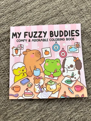 My Fuzzy Buddies målarbok - 24 sidor, några exempel på bilderna. En söt och färgglad målarbok med titeln 'My Fuzzy Buddies'. Omslaget har gulliga djurfigurer i pastellfärger och insidan bjuder på teckningar att färglägga, bland annat med björnar och andra djur runt en lägereld. Perfekt för dig som gillar kawaii och kreativa aktiviteter.