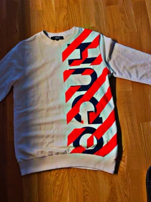 Vit HUGO sweatshirt med röd print  - Vit sweatshirt från HUGO med stora röda diagonala ränder och blå logotyptryck längs framsidan. Tröjan har rund halsringning och ribbade muddar vid ärmslut och nederkant. Perfekt för en trendig streetwear-look. Jätte fin äkta Hugo tröja riktigt fin  sitter bra på de mesta 