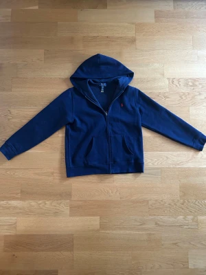 Mörkblå hoodie från Polo Ralph Lauren - Mörkblå hoodie med dragkedja från Polo Ralph Lauren. Klassisk design med huva. Det finns en QR länk på bild 3 som visar att varan är äkta. Storlek 170cm. Har endast haft på tröjan 1-3 gånger 