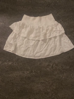 Vit volangkjol med smock i midjan - Söt vit kortkjol med bred smock i midjan och dubbla volanger. Perfekt för en somrig look och lätt att matcha med olika toppar. Kjolen har en luftig känsla och passar bra till varma dagar.