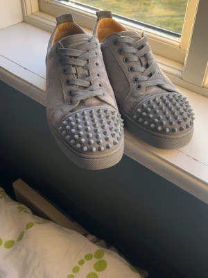Gråa Louboutin sneakers med nitar - Säljer ett par gråa sneakers från Christian Louboutin med ikoniska röda sulor och coola nitar på tån. Skorna är i mocka och har klassisk låg modell med matchande gråa skosnören. Perfekta för dig som vill sticka ut med en edgy stil.🔥
