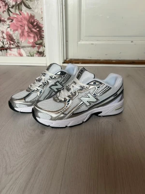 New Balance vita och silver sneakers - Snygga sneakers från New Balance i vitt och silver med mesh- och syntetmaterial. Klassisk chunky sula och tydliga loggor på sidan och plös. Skorna har snörning och sportig design som passar perfekt till streetwear och casual outfits.