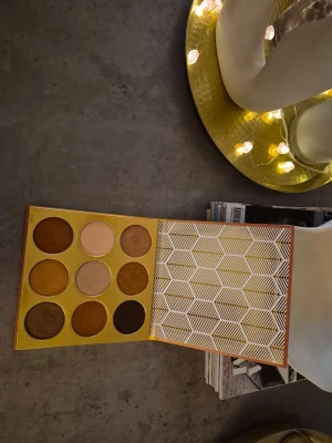 Ögonskuggspalett i varma toner - Snygg ögonskuggspalett med nio nyanser i bruna, guldiga och beige toner. Paletten har både matta och skimrande skuggor, perfekt för att skapa olika looks. Förpackningen är gul med geometriskt mönster på insidan. Skuggorna är pressade i runda pannor. The warrior by juvias. Paletten är swachad, testad, en fin palett 