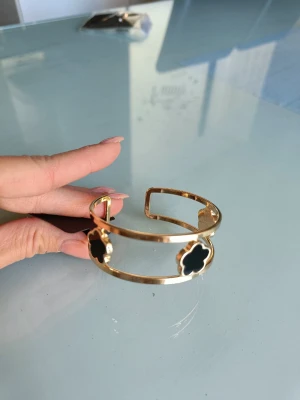 Guldfärgat armband med svarta detaljer - Snyggt guldfärgat armband med öppen design och tre fyrklöverformade svarta detaljer. Armbandet har en modern och stilren look som passar perfekt till många outfits. Tillverkat i metall med blanka svarta inslag i form av blommor.