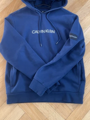 Blå Calvin Klein hoodie med tryck - Snygg blå hoodie från Calvin Klein Performance med vit logga på bröstet och ärmen. Klassisk huva med dragsko och känguruficka framtill. Mjuk insida och ribbade muddar. Perfekt för en chill och stilren look.