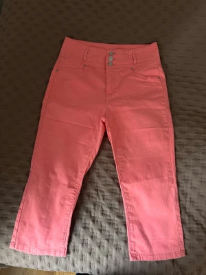 Rosa raka byxor med hög midja - Snygga rosa byxor med hög midja, trekvart långa och raka ben. Byxorna har tre knappar framtill, klassiska fickor och en bakficka med diskret broderi. Materialet känns mjukt och bekvämt.