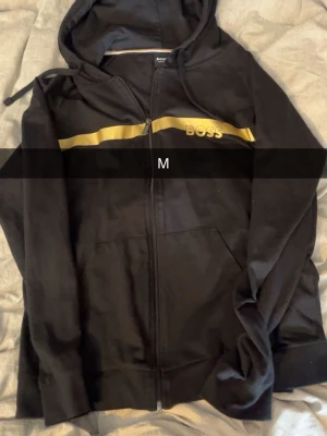Svart hoodie från BOSS med dragkedja - Snygg svart hoodie från BOSS med dragkedja framtill och huva med dragsko. Har en guldfärgad rand över bröstet och BOSS-logga på vänster sida. Perfekt för en stilren och sportig look.