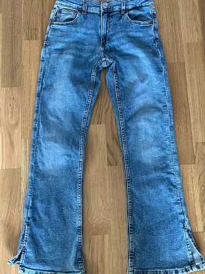 Blå bootcut jeans med slits nertill - Snygga ljusblå bootcut jeans med slits nertill på benen. Klassisk femficksmodell med dragkedja och knapp framtill. Jeansen har en avslappnad passform och är tillverkade i denim. Perfekta för en trendig och avslappnad look.