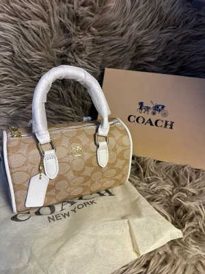 Beige och vit handväska - Snygg handväsk i beige canvas  och vita detaljer i läder. Väskan har guldfärgade metalldetaljer, dubbla handtag och dragkedja upptill. Perfekt accessoar för att lyfta din outfit.