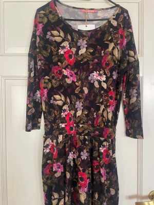 Blommig långklänning med 3/4-ärm - Klänning från Pernilla Wahlgren med vackert blommigt mönster i rött, rosa, lila och beige på mörk botten. Klänningen har 3/4-ärmar och en avslappnad passform med markerad midja. Perfekt för dig som gillar färg och mönster!