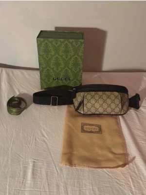 Gucci beige bälte-väska med monogram - Snygg beige bälte-väska från Gucci med klassiskt GG-monogram och svarta detaljer. Väskan har justerbart svart bälte, dragkedja och extra ficka framtill. Kommer med originalförpackning och dustbag. Perfekt för att bära det viktigaste med stil.