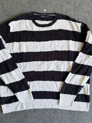 Svartvit kabelstickad tröja Nautica - Snygg kabelstickad tröja från Nautica i storlek L med breda svarta och vita ränder. Tröjan har rund halsringning, ribbade muddar och är perfekt för dig som gillar klassisk och sportig stil. Materialet är mjukt och bekvämt.