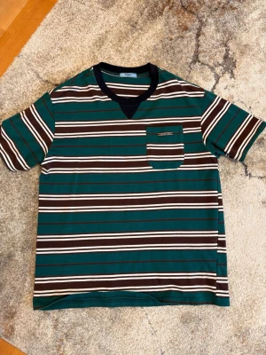 Prada randig t-shirt herr - Randig t-shirt från Prada i bomull, stl XL, med gröna, bruna och vita ränder. Kort ärm, rund hals med mörk kant och bröstficka med liten logodetalj.
