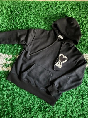 Svart hoodie från Ami Paris med hjärtdetalj - Svart hoodie från Ami Paris med stor vit hjärtdetalj och bokstaven A på bröstet. Klassisk känguruficka framtill, ribbade muddar och dragsko i huvan. Mjuk bomullskänsla och stilren design, perfekt för en trendig streetwear-look.