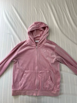 Rosa velourhoodie från Juicy Couture - Mjuk rosa hoodie i velour från Juicy Couture med dragkedja framtill och stor känguruficka. Klassisk huva och diskret logga broderad på bröstet. Perfekt för en chill och trendig look.