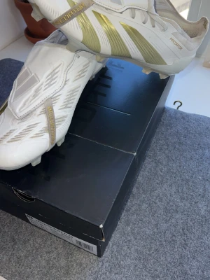 Adidas Predator fotbollsskor elite  - Snygga Adidas Predator fotbollsskor i vitt med guldiga detaljer och klassiska stripes. Skorna har låg profil, snörning och täckt plös för bättre bollkontroll. Tillverkade i syntetmaterial och designade för spel på gräsplan. Perfekta för dig som vill sticka ut på planen.