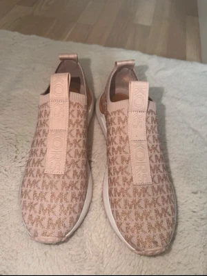 Rosa slip-on sneakers från Michael Kors - Snygga slip-on sneakers från Michael Kors i ljusrosa med MK-logga över hela ovandelen. Skorna har bred resår med logotyp framtill och vit sula. Tillverkade i textil med stickad känsla och elastisk kant runt ankeln för extra komfort. Använd 1 gång