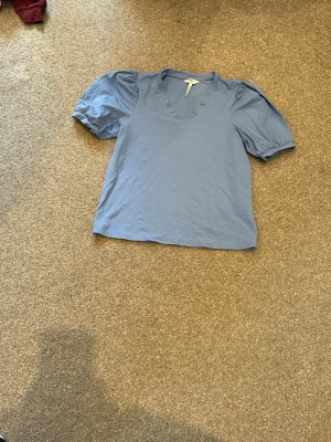 Blå topp med puffärm och v-ringning - Snygg blå topp med korta puffärmar och v-ringning. Perfekt för dig som gillar en enkel men trendig stil. Toppens material känns mjukt och bekvämt mot huden. Passar till både jeans och kjol för en avslappnad look.