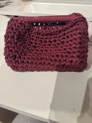 vinröd clutchbag, handvrikad  - Unik handvirkad kuvertväska i vinrött garn med grov struktur. Väskan har ett inbyggt handtag upptill och är rektangulär i formen. Perfekt accessoar för dig som vill sticka ut med en personlig touch. Materialet är mjukt och flexibelt tyg. Jag har virkat den själv med material från panduro. Mått: ca 27x17x15cm