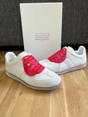 Maison Margiela ”Japan” - Maison Margiela sneakers i vitt läder med röda skosnören och röd sprayad detalj på sidan. Klassisk låg modell med grå sula och paneler i mocka framtill. Snygg och unik design som sticker ut.