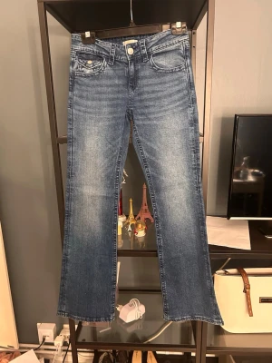 Blå bootcut jeans med broderade fickor - Säljer ett par blå bootcut jeans med snygg tvätt och broderade bakfickor med rosettdetaljer. Jeansen har klassisk femficksmodell, knappgylf och raka ben. Perfekta för dig som gillar en avslappnad men ändå trendig stil.