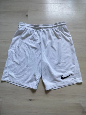 Vita sportshorts från Nike - Säljer ett par klassiska vita sportshorts från Nike med elastisk midja och svart logga på ena benet. Perfekta för träning eller fotboll. Lätta och bekväma i syntetmaterial som andas.