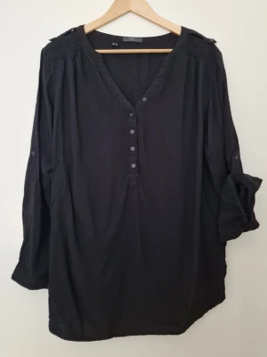 Svart blus med knappar från BPC Collection  - Stilren svart blus från Esmara med v-ringning och dekorativa knappar framtill. Blusen har långa ärmar som kan rullas upp och fästas med knapp. Tillverkad i ett lätt och luftigt tyg, perfekt för en avslappnad men snygg look. Storlek 40. Fint skick. 
