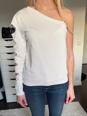 Vit off-shoulder tröja med cut-outs - Trendig vit off-shoulder tröja med långa ärmar och unika cut-outs längs ena ärmen. Tröjan har en avslappnad passform och ribbade muddar vid ärmslut och nederkant. 