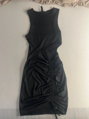 Svart kort klänning med snörning - Snygg svart kort klänning i figurnära modell med ärmlös design och cool snörningsdetalj framtill. Klänningen har en cut-out vid midjan och är tillverkad i ett glansigt syntetmaterial som ger en modern look.