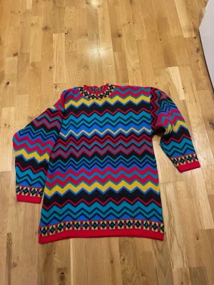 Färgglad zigzag-stickad tröja stl M - Säljer en långärmad stickad tröja med färgstarkt zigzag-mönster i blått, rött, gult och svart. Tröjan har ribbade muddar och detaljerade kanter med småmönstrat motiv. Perfekt för dig som gillar unika och lekfulla plagg!