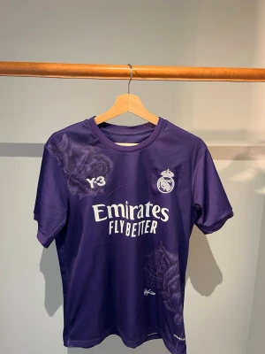 Real Madrid - Snygg lila matchtröja från Real Madrid i samarbete med Y-3. Tröjan har vita tryck med klubbmärke, sponsorlogga och Y-3 på bröstet samt diskreta blommönster. Tillverkad i lätt och funktionellt material, perfekt för fotboll eller träning.
