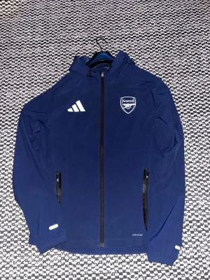 Mörkblå Arsenal träningsjacka Tiro 25 Adidas Competition - Säljer en Mörkblå träningsjacka från Adidas med Arsenal-logga på bröstet. Jackan har huva, hel dragkedja, två fickor med dragkedja och vita detaljer. Tillverkad i lätt och funktionellt material med Aeroready-teknologi. Kan byta mot Manchester united Tiro25