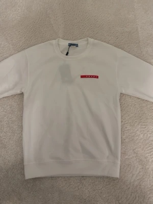 Vit sweatshirt från Prada - Säljer en clean vit sweatshirt från Prada med klassisk rund hals och ribbade muddar. På bröstet sitter en röd logotyp med PRADA-tryck som ger en exklusiv vibe. Perfekt för dig som gillar minimalistisk och stilren design. Materialet känns mjukt och bekvämt.