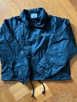 Svart vintage vindjacka från Champion windbreaker  - Snygg svart vindjacka från Champion med klassisk krage och dragsko i nederkant. Jackan har tryckknappar framtill och logga broderad på bröstet. Perfekt för blåsiga dagar och enkel att matcha med olika outfits.