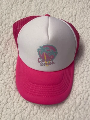 Rosa och vit truckerkeps Coco Beach - Snygg truckerkeps i rosa och vitt med tryck av palmer och texten 'Coco Beach' framtill. Kepsen har böjd skärm, mesh på sidorna och justerbar snapback. Perfekt för en avslappnad och cool stil.