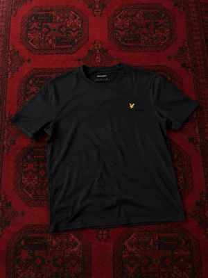 Svart t-shirt från Lyle & Scott - Svart t-shirt från Lyle & Scott med klassisk passform och rund hals. T-shirten har en gul broderad logotyp på bröstet och är tillverkad i mjuk bomull. Perfekt basplagg med stilren look.